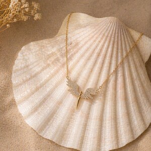 May include: A gold-colored necklace with a pendant shaped like angel wings, encrusted with small, clear crystals. The necklace is displayed on a large, cream-colored seashell, set against a sandy background. Delicate dried flowers are visible in the upper left corner.