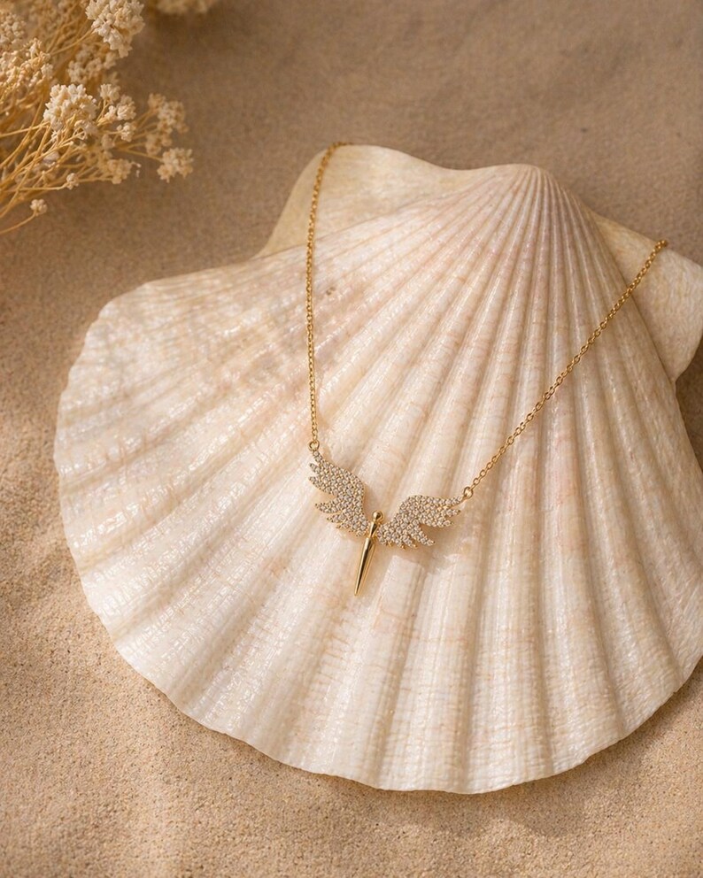 May include: A gold-colored necklace with a pendant shaped like angel wings, encrusted with small, clear crystals. The necklace is displayed on a large, cream-colored seashell, set against a sandy background. Delicate dried flowers are visible in the upper left corner.
