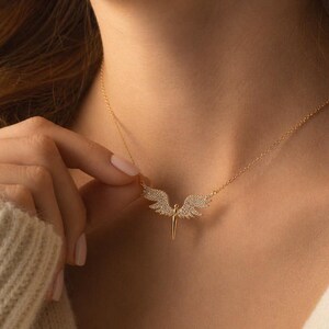 May include: A gold-colored necklace featuring a pendant shaped like angel wings. The wings are encrusted with small, sparkling stones. The necklace is displayed on a person's neck, with the chain visible.