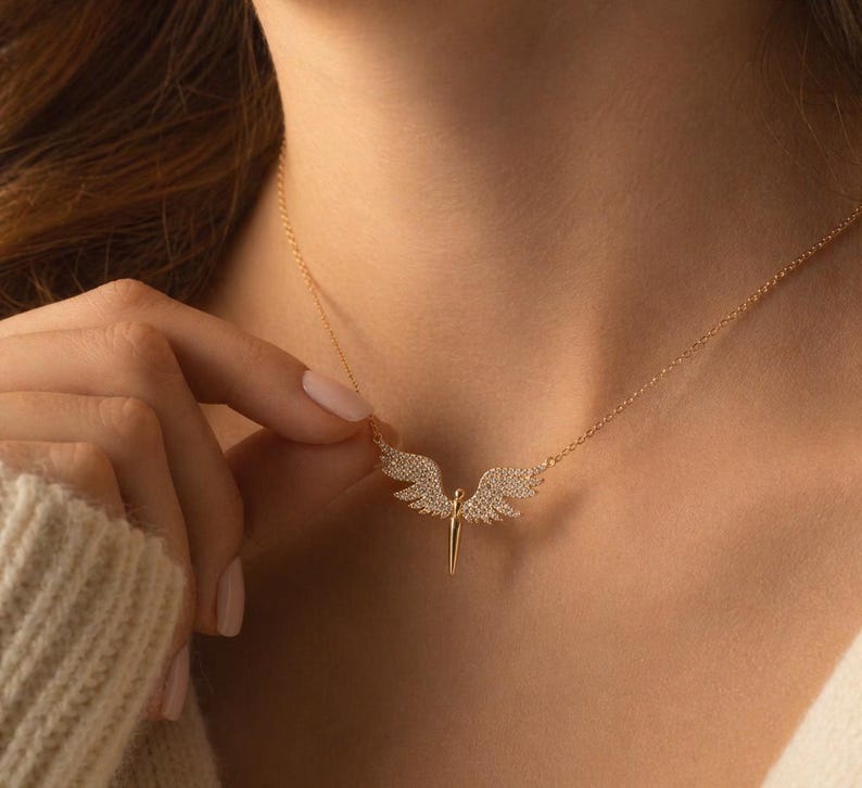 May include: A gold-colored necklace featuring a pendant shaped like angel wings. The wings are encrusted with small, sparkling stones. The necklace is displayed on a person's neck, with the chain visible.