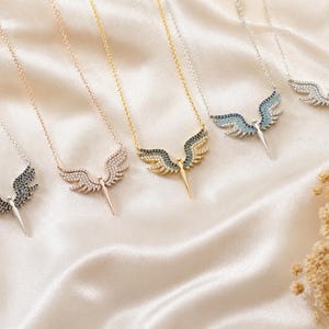 May include: A collection of necklaces with angel wing pendants. The pendants feature various colors of gemstones, including black, clear, and blue, set in silver, gold, and rose gold tones. The necklaces rest on a cream-colored satin surface.