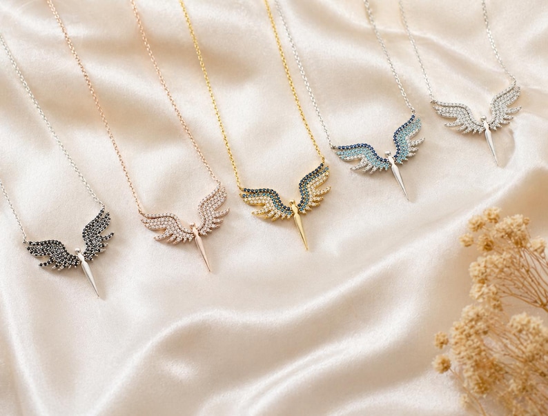 May include: A collection of necklaces with angel wing pendants. The pendants feature various colors of gemstones, including black, clear, and blue, set in silver, gold, and rose gold tones. The necklaces rest on a cream-colored satin surface.