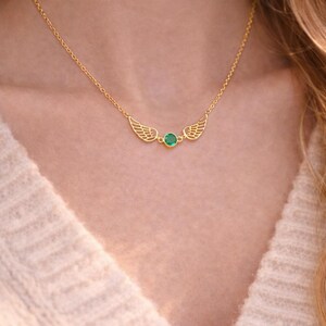 May include: A delicate gold-colored necklace featuring a pair of stylized wings flanking a small, round, green gemstone. The necklace is worn against a cream-colored sweater, highlighting the jewelry's design.