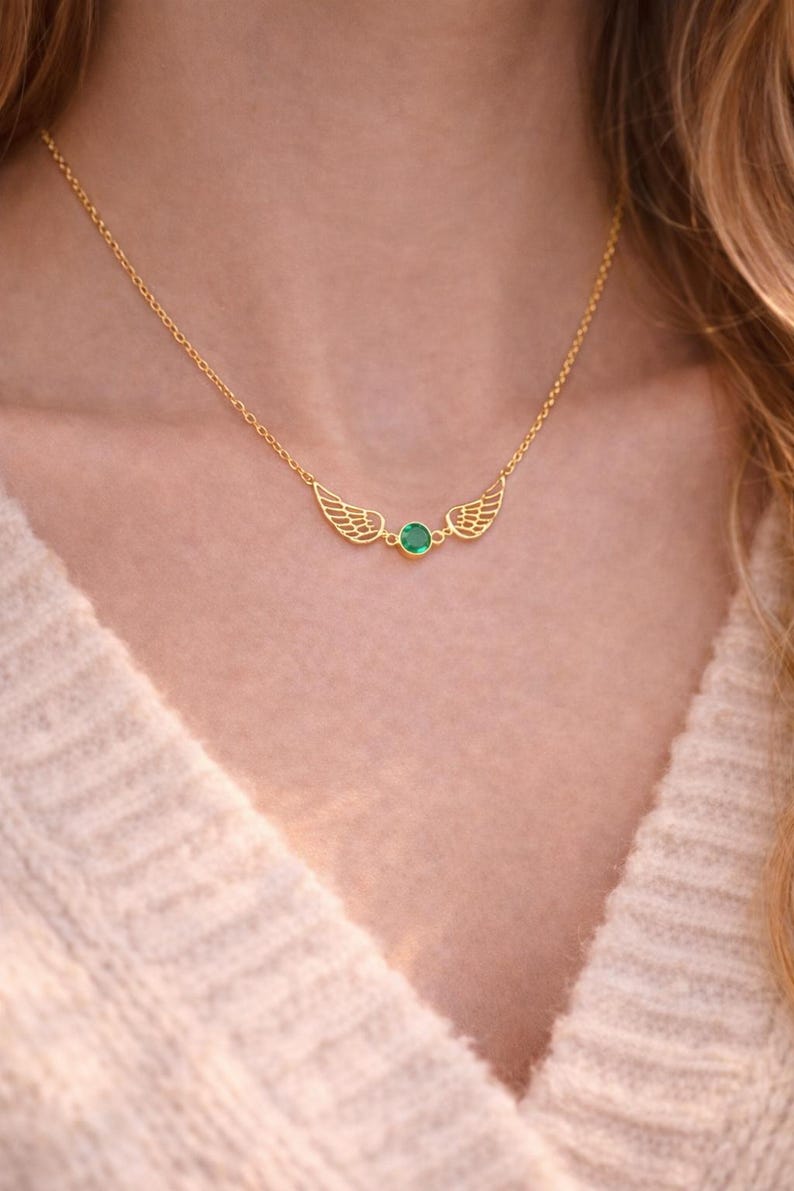 May include: A delicate gold-colored necklace featuring a pair of stylized wings flanking a small, round, green gemstone. The necklace is worn against a cream-colored sweater, highlighting the jewelry's design.