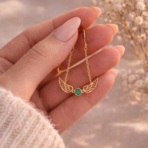 May include: Gold-colored earrings featuring a wing design with a small, round, green gemstone in the center. The earrings are suspended from delicate chains. The jewelry is displayed against a soft, neutral background.