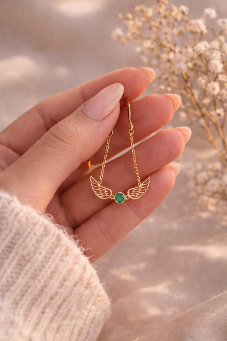 May include: Gold-colored earrings featuring a wing design with a small, round, green gemstone in the center. The earrings are suspended from delicate chains. The jewelry is displayed against a soft, neutral background.