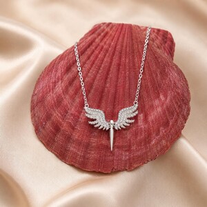 May include: A silver-toned necklace featuring a pendant shaped like angel wings with a central, pointed element. The wings and pendant are encrusted with small, sparkling stones. The necklace rests on a red seashell against a satin background.