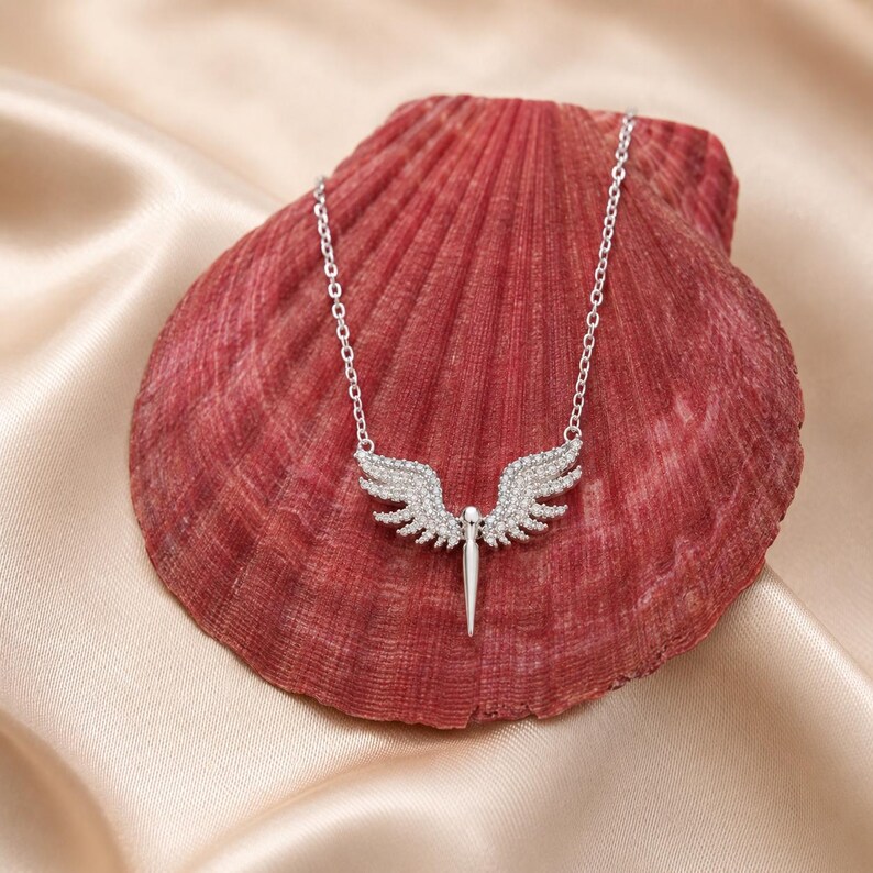 May include: A silver-toned necklace featuring a pendant shaped like angel wings with a central, pointed element. The wings and pendant are encrusted with small, sparkling stones. The necklace rests on a red seashell against a satin background.