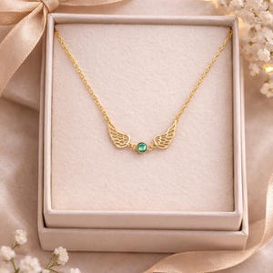 May include: A gold-colored necklace with a delicate chain and wing-shaped pendants on either side of a small, round, green gemstone. The necklace is presented in a white gift box with a satin ribbon bow.