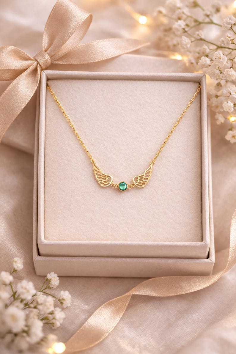 May include: A gold-colored necklace with a delicate chain and wing-shaped pendants on either side of a small, round, green gemstone. The necklace is presented in a white gift box with a satin ribbon bow.