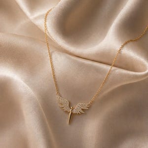May include: A gold-colored necklace with a pendant shaped like angel wings. The wings are encrusted with small, clear gemstones. The necklace is displayed on a beige satin fabric, creating a luxurious aesthetic. The pendant has a central, vertical spike.