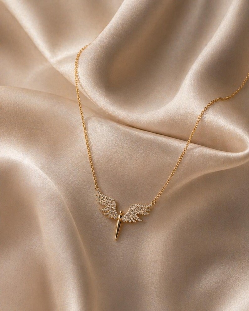 May include: A gold-colored necklace with a pendant shaped like angel wings. The wings are encrusted with small, clear gemstones. The necklace is displayed on a beige satin fabric, creating a luxurious aesthetic. The pendant has a central, vertical spike.