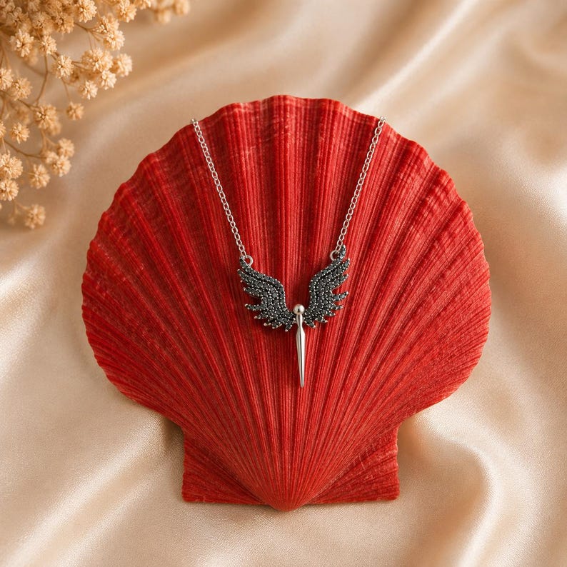 May include: A silver-toned necklace featuring a pendant with black gemstone-studded wings and a central, elongated silver element. The necklace rests on a vibrant red seashell against a cream-colored satin backdrop. The jewelry design is elegant.