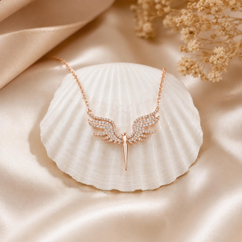 May include: Rose gold-toned necklace featuring a pendant shaped like angel wings. The wings are encrusted with small, sparkling crystals. The necklace rests on a white seashell against a backdrop of soft, cream-colored fabric.