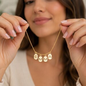 May include: A gold-colored necklace with the name "EMMA" spelled out in individual letter pendants. The letters are in a simple, sans-serif font and are suspended from a delicate chain. The necklace is being held up by a person.