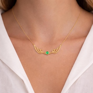 May include: A gold-colored necklace featuring a pair of openwork wings on either side of a round, green gemstone. The necklace is delicate and appears to be made of a thin chain.
