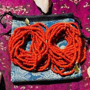 May include: Two sets of orange beaded necklaces, each with a gold clasp, are displayed on a blue and white patterned fabric pouch. The pouch is on a purple fabric background.