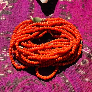May include: A long strand of bright orange coral beads with a gold leaf charm. The beads are arranged in a circular shape on a purple and white patterned fabric.