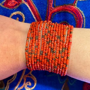 May include: A stack of orange and gold beaded bracelets on a person's wrist. The bracelets are made of small, round beads and are worn on a blue background.