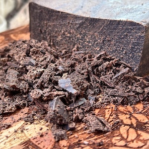 May include: A close-up of a dark chocolate block with a rough, textured surface. Chocolate shavings are scattered in front of the block on a wooden surface with carved floral designs. The chocolate appears rich and decadent.
