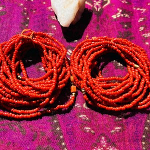 May include: Two strands of red beaded jewelry, each wrapped in a circle, on a purple fabric background.