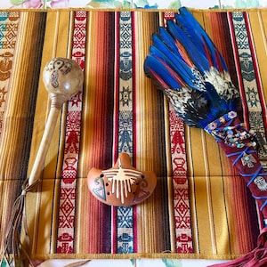 Puede incluir: Un surtido de artículos indígenas hechos a mano, que incluyen una sonaja de calabaza, una ocarina de cerámica y un abanico de plumas con detalles azules y rojos. Los artículos se exhiben sobre un textil a rayas con patrones geométricos.