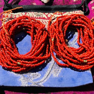 May include: Two strands of red coral beads, each with gold accents, are wrapped around a blue and silver patterned fabric pouch.