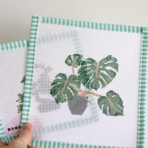 Puede incluir: Dos lienzos cuadrados de punto de aguja con una planta de Monstera verde en una maceta gris. El punto de aguja está sobre lienzo blanco con un borde a rayas verdes y blancas. La planta tiene hojas verdes grandes y tallos marrones.