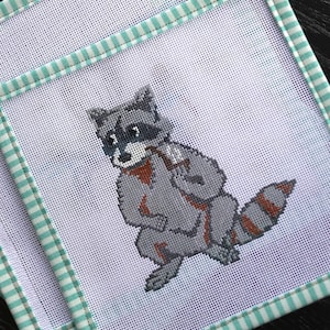 May include: Two white canvas needlepoint kits with a green and white striped border. The kits feature a raccoon design.