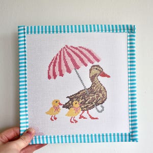 Puede incluir: Un lienzo de punto de aguja con un borde a rayas azul y blanco. El lienzo presenta un pato marrón con dos patitos amarillos. El pato sostiene un paraguas a rayas rosa y blanco.