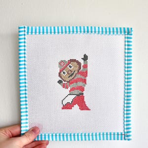 May include: A white canvas needlepoint with a blue and white striped border. The canvas features a cross-stitch design of a cartoon mascot wearing a red and white striped shirt and red pants.