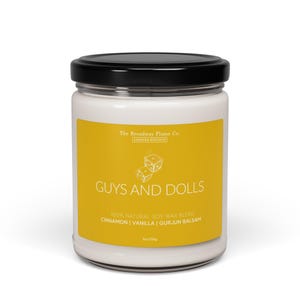 Könnte beinhalten: Ein gelbes Kerzengefäß mit schwarzem Deckel. Das Etikett lautet "The Broadway Flame Co. Limited Edition" und "Guys and Dolls". Das Etikett zeigt auch "100% Natural Soy Wax Blend" und "Cinnamon | Vanilla | Gurjun Balsam".