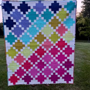 May include: A colorful quilt with a geometric pattern of intersecting squares in shades of blue, teal, green, pink, and purple. The quilt is held up against a backdrop of green grass and a soft, dusky sky.