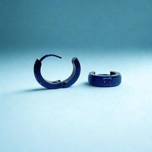 May include: Two dark blue hoop earrings. One earring is open, showing the clasp, while the other is closed. The earrings have a textured surface and a simple, modern design. The background is a solid light blue.