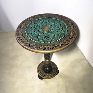 May include: A round, decorative side table with a detailed, ornate design. The tabletop features a teal and gold mandala pattern, with a black and gold border. The table has a single pedestal base and measures approximately 24 inches in diameter.