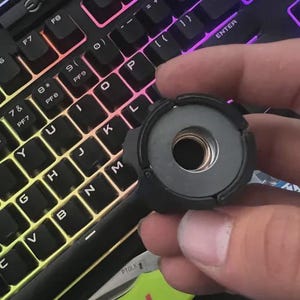 May include: A black, circular object with a threaded center is held in front of a backlit keyboard. The object has a handle extending from it. The keyboard keys are black with white lettering and have a rainbow glow.
