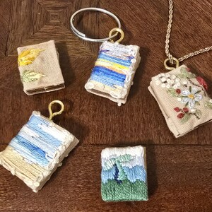 May include: Five small embroidered fabric books with different designs. The books are beige with white stitching and feature various colorful embroidery designs, including a sunset, clouds, a mountain, and flowers.