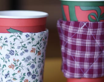 May include: Several coffee cups with decorative fabric sleeves. One sleeve features a floral pattern, another a plaid design. The cups are red and green, with white lids. A matching floral coaster is also visible.