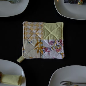 May include: A quilted square pot holder with a loop, featuring a patchwork design of yellow and floral fabrics. The pot holder is surrounded by white plates, forks, and a napkin ring on a black surface.