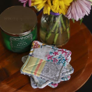 May include: A green candle with a silver lid and the words "WHITE BARN" and "FRESH BALSAM" printed on the label. A stack of patchwork coasters with floral and geometric patterns sits on a wooden tray, next to a vase of flowers.