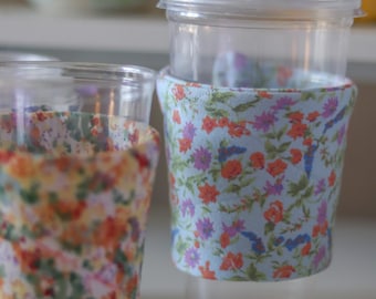 May include: Two clear plastic cups with floral fabric sleeves. One sleeve has a blue background with orange, purple, and green flowers. The other sleeve has a yellow background with orange, red, and green flowers. One cup has a clear plastic lid.
