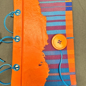 Peut inclure: Un journal fait à la main avec une couverture en cuir orange et un dos en tissu coloré. Le journal est relié avec un cordon bleu et comporte une fermeture à bouton orange. Le dos en tissu présente des rayures bleues, violettes et oranges.