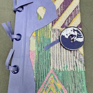 Peut inclure: Un journal fait à la main avec une couverture en cuir lavande et un motif en tissu tissé coloré. La couverture présente une découpe unique et est fixée avec des liens en cuir. Un bouton bleu marine avec un motif blanc orne le tissu.