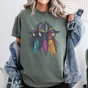 Pode incluir: Camiseta verde-oliva com uma ilustração em aquarela de três bruxas usando chapéus pontudos. As bruxas têm cabelos longos em tons de azul, roxo e vermelho. O texto "Pure Grit Designs" é visível no canto inferior esquerdo.