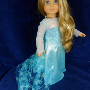 May include: A doll with long blonde hair and blue eyes, wearing a light blue dress with a snowflake design. The dress has a sheer overlay and a long train. The doll has snowflake hair accessories.