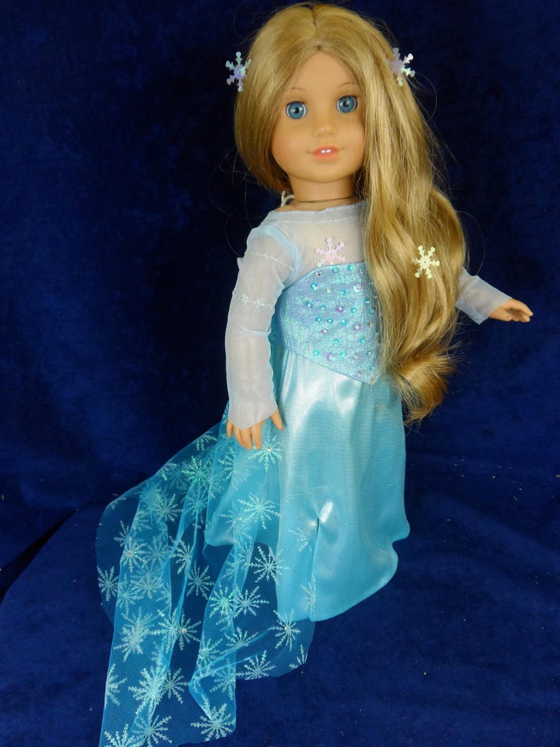 May include: A doll with long blonde hair and blue eyes, wearing a light blue dress with a snowflake design. The dress has a sheer overlay and a long train. The doll has snowflake hair accessories.
