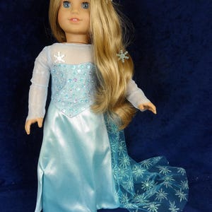 May include: A doll with long blonde hair and blue eyes, wearing a light blue dress with a snowflake design. The dress has a sheer overlay and a long, flowing skirt. Snowflake hair accessories complete the look.