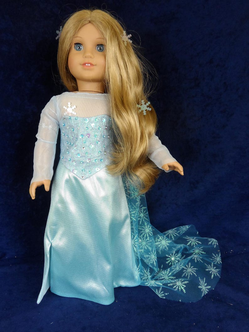 May include: A doll with long blonde hair and blue eyes, wearing a light blue dress with a snowflake design. The dress has a sheer overlay and a long, flowing skirt. Snowflake hair accessories complete the look.