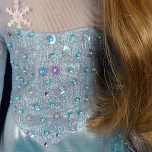 May include: A light blue doll dress with a bodice embellished with iridescent sequins and beads. The dress features a sheer neckline and a snowflake applique. Long blonde hair is visible on the right side of the image.