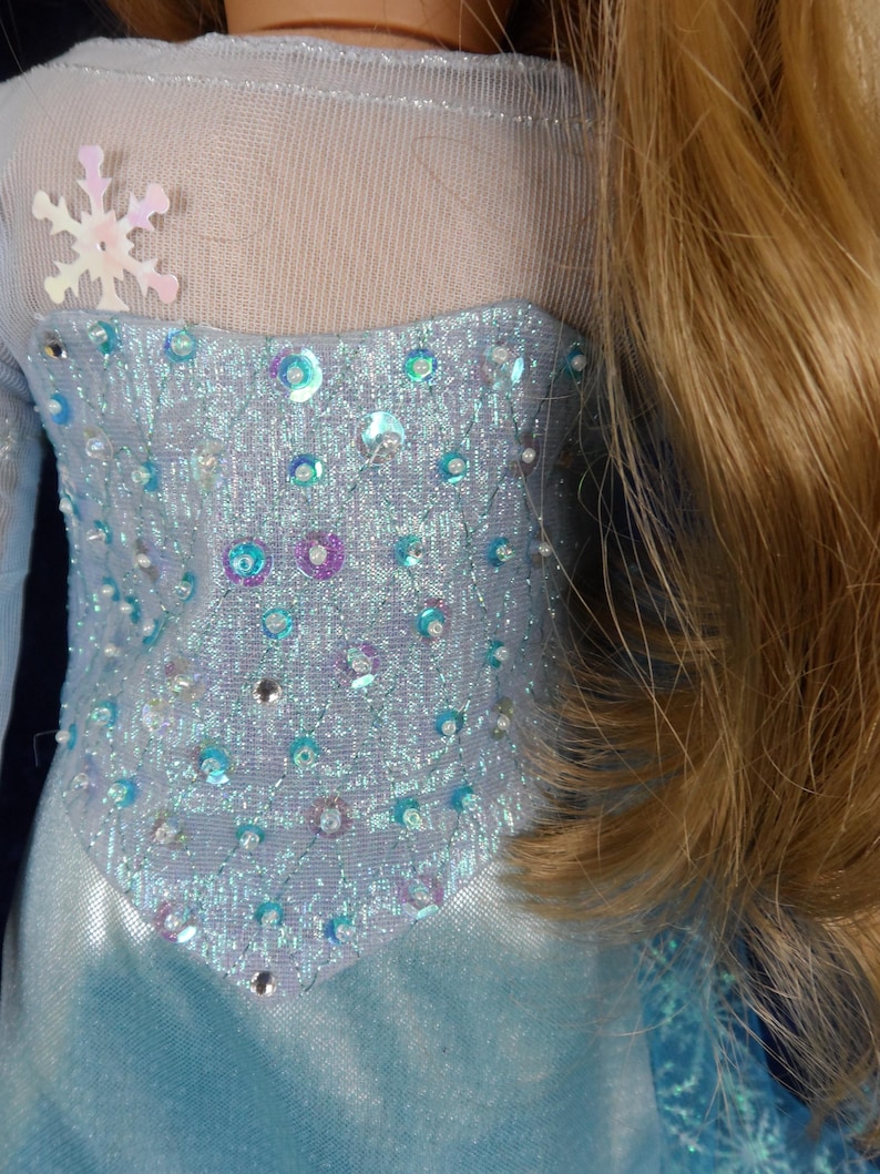 May include: A light blue doll dress with a bodice embellished with iridescent sequins and beads. The dress features a sheer neckline and a snowflake applique. Long blonde hair is visible on the right side of the image.